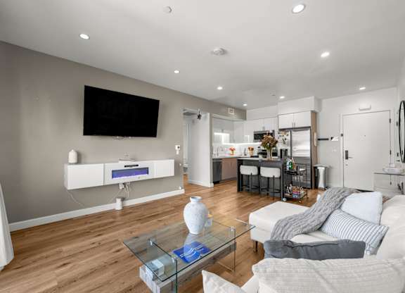 A modern living room with a white sofa, a glass coffee table, and a flat-screen TV mounted on the wall.