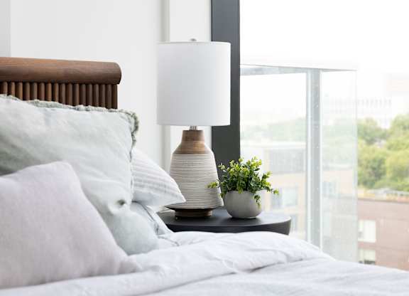 A bed with white pillows and a grey blanket with a lamp and a plant on the bedside table.