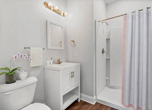 A white bathroom with a white tub, a white cabinet, a white toilet, a white sink, a white mirror, a white towel, a white vase with purple flowers, a white tray with a soap dish