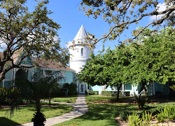 A white building with a tower is surrounded by green trees.