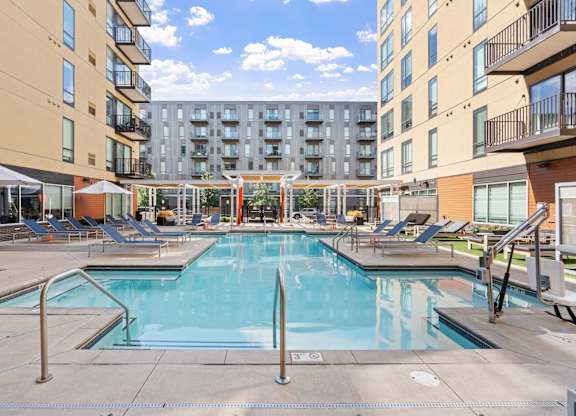 A large swimming pool surrounded by lounge chairs and umbrellas in a sunny courtyard.