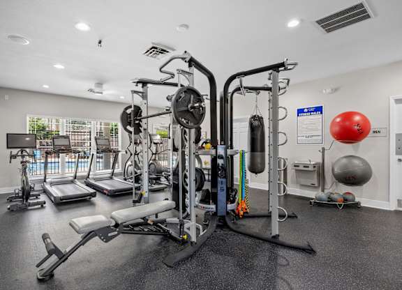 A gym with a variety of equipment including a weight bench, a leg press machine, a cable machine, a treadmill, a stationary bike, a rowing machine, a medicine ball, and a punching bag.