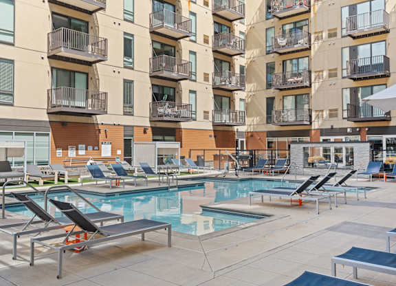 A large outdoor pool area with sun loungers and apartment buildings in the background.