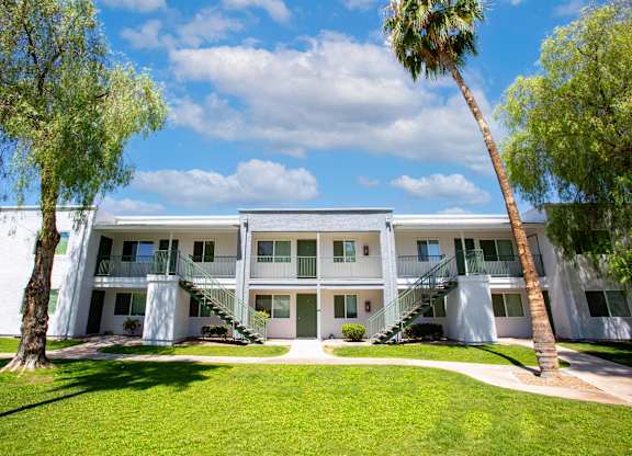 A white building with a balcony on the second floor is surrounded by green grass and trees.