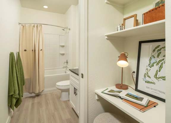 A bathroom with a toilet, a shower with a curtain, a shelf with a lamp and a picture frame.