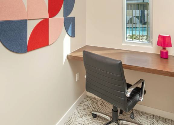 A red, blue and grey abstract art piece hangs on a wall above a desk with a black chair.