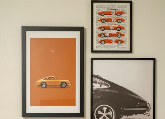 Three framed pictures of cars are hanging on a wall above a bed with a white comforter and two pillows.
