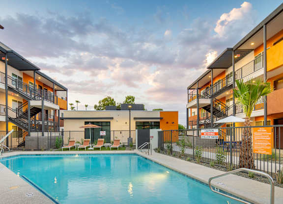 A swimming pool is surrounded by a fence and chairs in front of a building.