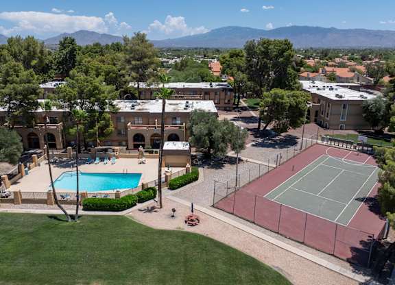 A tennis court is surrounded by a fence and a pool is in the middle of the grassy area.