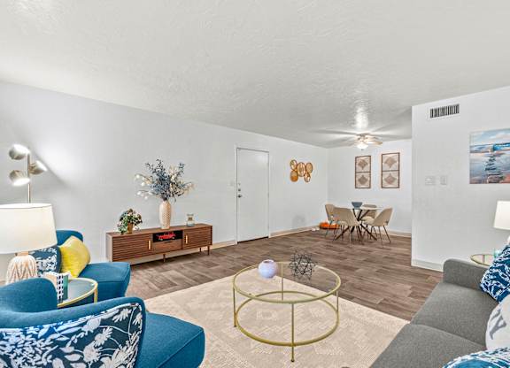 A living room with a grey sofa, a blue armchair, a wooden side table with a vase of flowers, a coffee table with a gold base, a wooden cabinet with a mirror, a dining table with chairs, and