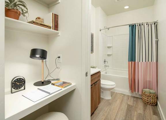A bathroom with a toilet, a sink, a shelf, a clock, a potted plant, a lamp, a towel rack, a basket, and a shower curtain.