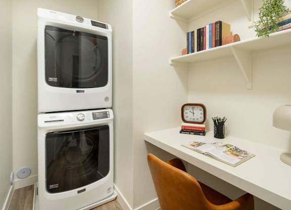 A white microwave oven is placed next to a white desk with a chair and a clock on it.