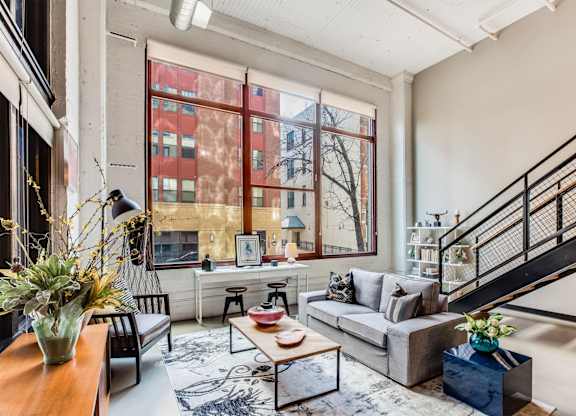 A living room with a grey couch, a coffee table, a staircase, and a large window.