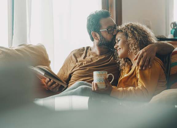 A man and woman are sitting on a couch, with the man holding a book and the woman holding a mug.