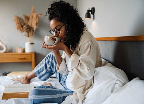 A woman in pajamas is sitting on a bed with a cup in her hand.