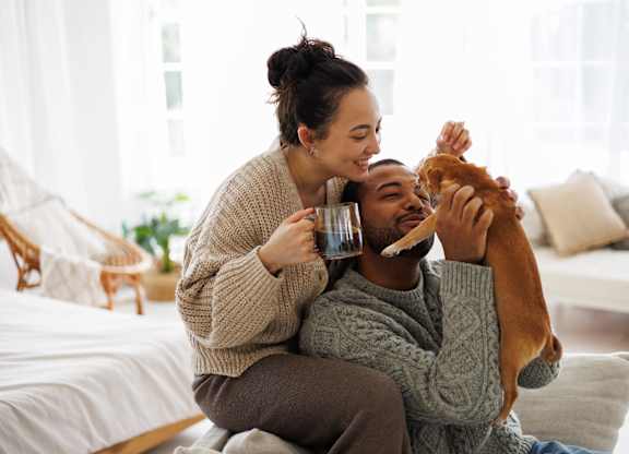 A woman is feeding a dog while sitting on a bed with a man.