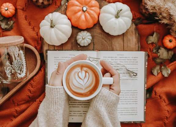 A person in a white sweater is holding a cup of coffee and reading a book.