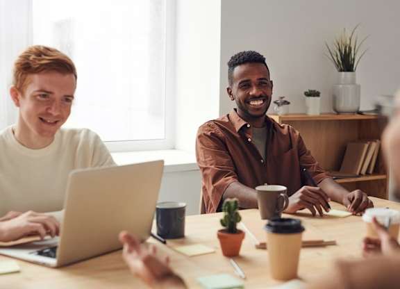 Three people are sitting around a table with a laptop, coffee cups, and a cactus plant.