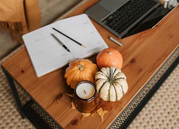 A wooden table with a laptop, a notebook, a pen, a pumpkin, and a candle on it.