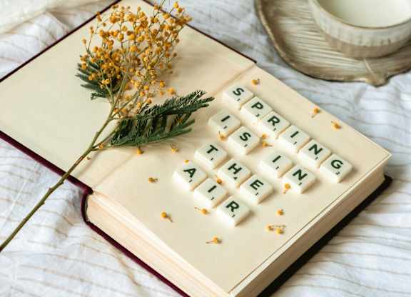 A book with the title "Spring is the Time" is open and placed on a bed with a cup of coffee and a jug of milk beside it.