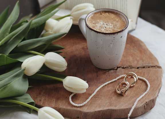 A cup of coffee and a bouquet of white tulips on a wooden board.