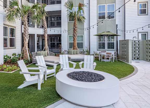 A white fire pit sits in the middle of a grassy area with white chairs around it.