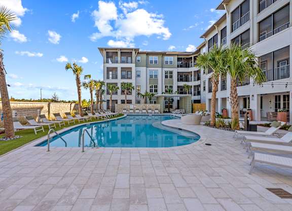 A large swimming pool surrounded by palm trees and lounge chairs in front of a multi-story apartment building.