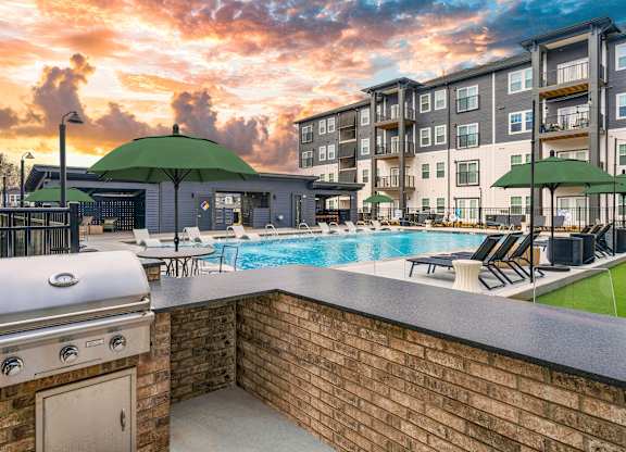 A sunset view of a pool area with a barbecue grill and lounge chairs.