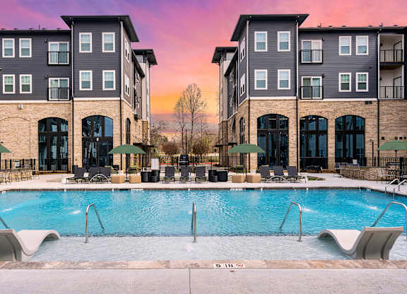 A swimming pool is surrounded by lounge chairs in front of apartment buildings.