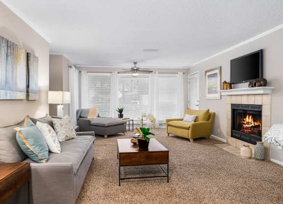 A carpeted living room with a tiel-surround fireplace, floor-to-ceiling windows, and a ceiling fan at Summerchase at Riverchase in Hoover, Alabama.