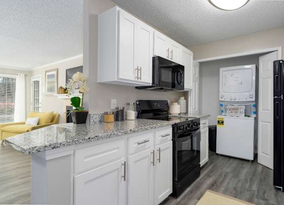 A galley-style kitchen with white cabinets, black appliances, granite countertops, and a washer and dryer in a laundry closet at Summerchase at Riverchase in Hoover, Alabama.