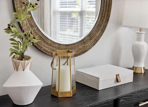 A white vase with a plant in it sits on a table next to a mirror and a white lamp.
