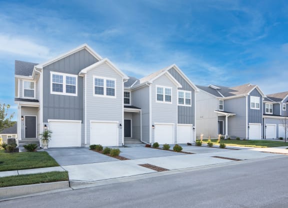 Raymore, MO, Townhomes Near Timbermist Farm - The Venue Townhomes - Exterior View of Townhomes with Grey-Toned Walls, White Trim, Parking Garage, Driveway, Manicured Lawns, Bushes, Trees, and Concrete Sidewalk.