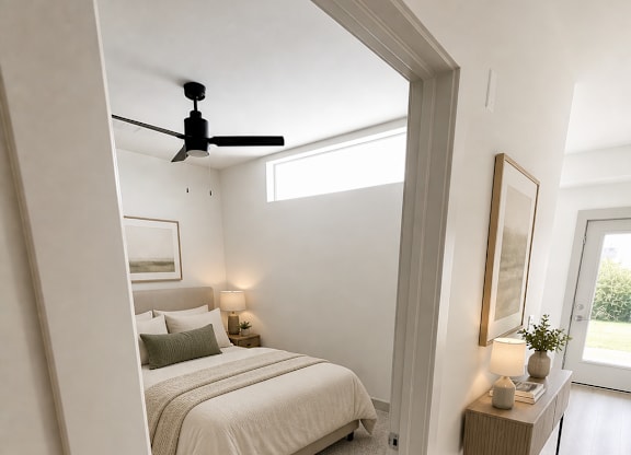 Pet-Friendly Apartments in Kokomo, IN - The Elwood Apartments - Bedroom With A Neutral Bed, A Ceiling Fan, High Window Light, And A Hallway Console Table With Lamp.