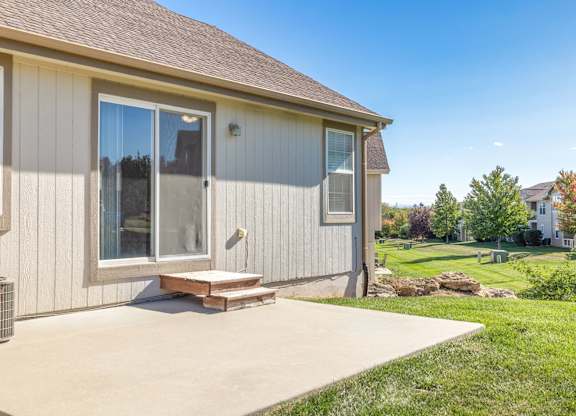 A house with a grey siding and a brown roof has a patio with a bench and a window.