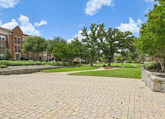A brick pathway leads to a grassy area with trees and a building in the background.