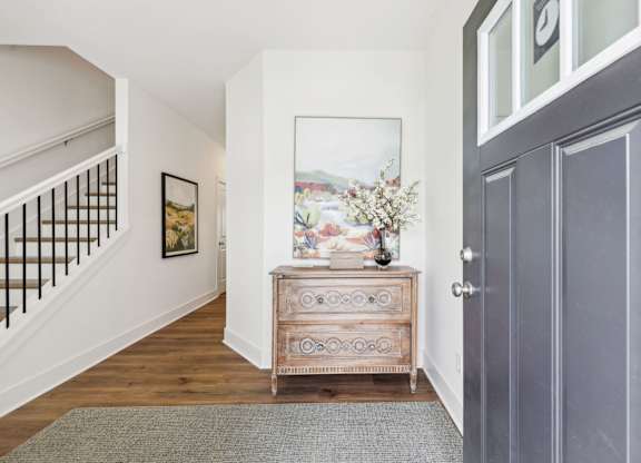 A hallway with a wooden floor, a grey rug, a wooden chest of drawers, a painting, and a staircase with a metal railing.