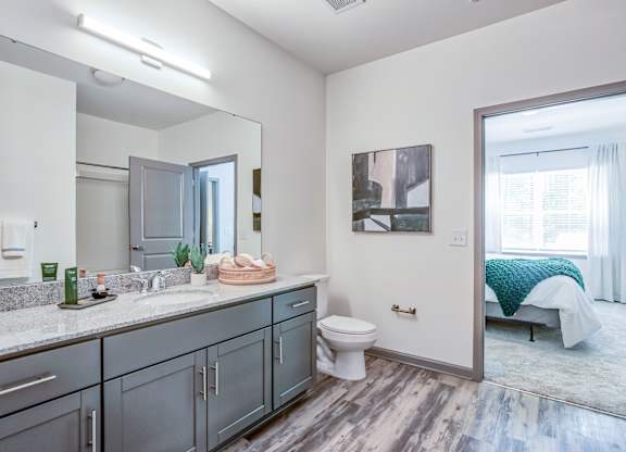 A bathroom with a sink, mirror, toilet, and a bedroom in the background.