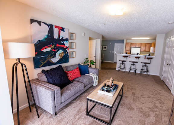 A grey couch with red and blue pillows sits in a living room with a painting on the wall.