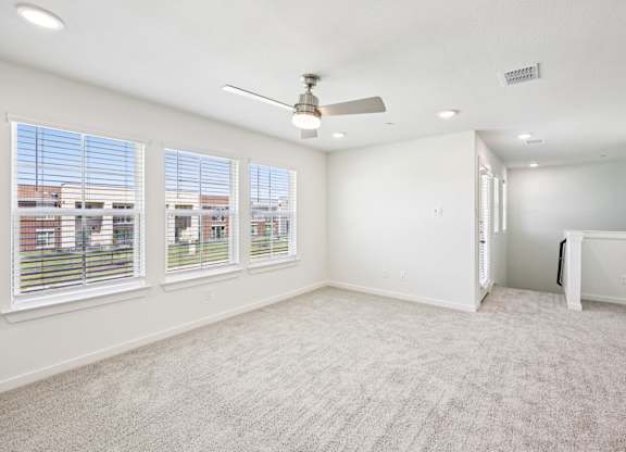 A room with a carpeted floor, a ceiling fan, and a view of a building outside the window.