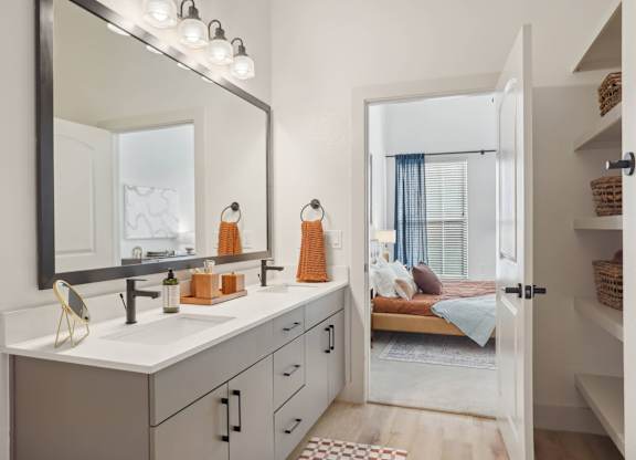 A bathroom with a vanity, mirror, and a bedroom in the background.