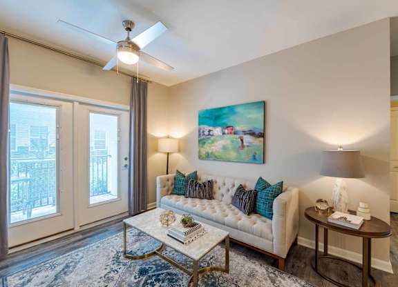A living room with a couch, a painting, a ceiling fan, and a table with a lamp.
