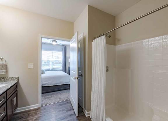 A white bathroom with a shower and a bedroom visible through the door.