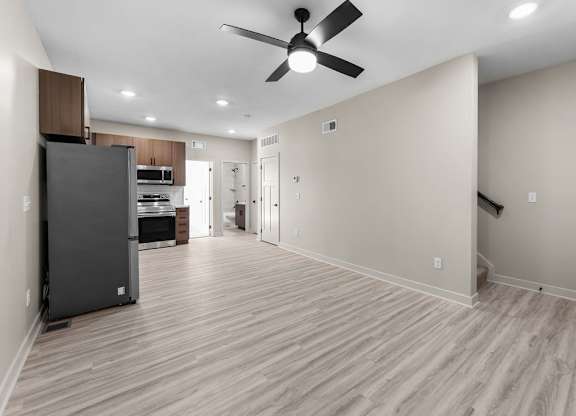 A spacious room with a ceiling fan and light, hardwood floors, and a kitchen area with a refrigerator.