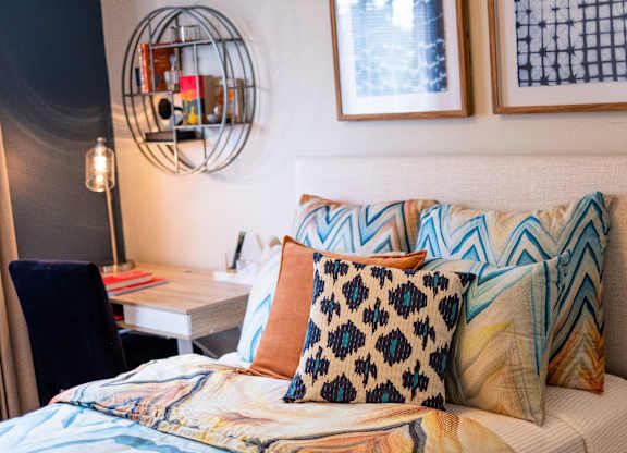 A bed with a blue and orange pillow is in the foreground of a room with a desk and chair.