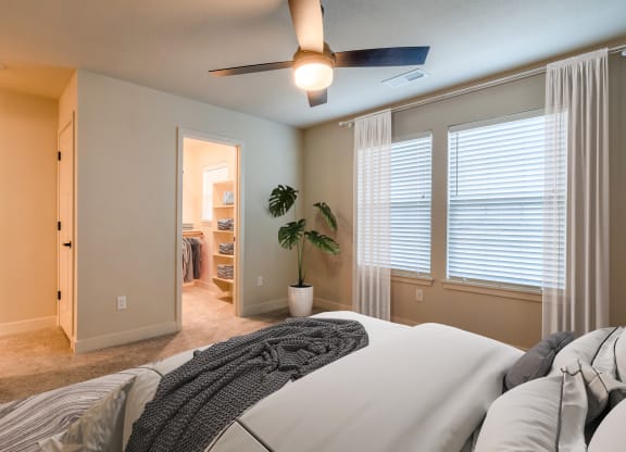Raymore, MO Townhomes - Bedroom With Ceiling Fan, Windows, Large Walk-In Closet, Carpet Flooring, And Two Windows.