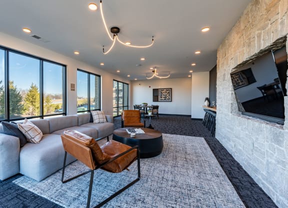 Carmel, IN, Apartments Near Monon Trail - North End - Clubroom with Carpeted Flooring, Couch, Armchairs, Coffee Table, TV, Kitchen Area, and Floor-to-Ceiling Windows.