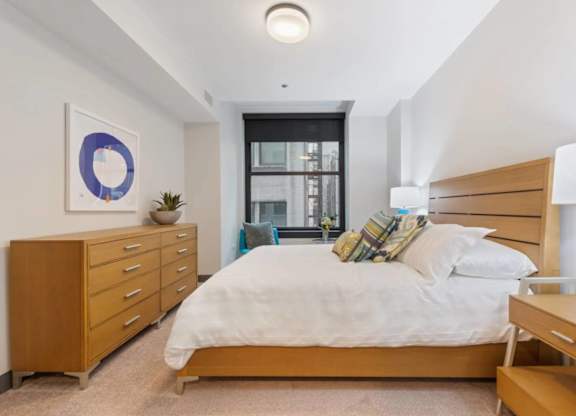A bedroom with a large bed, a wooden dresser, and a window with a view of a cityscape.