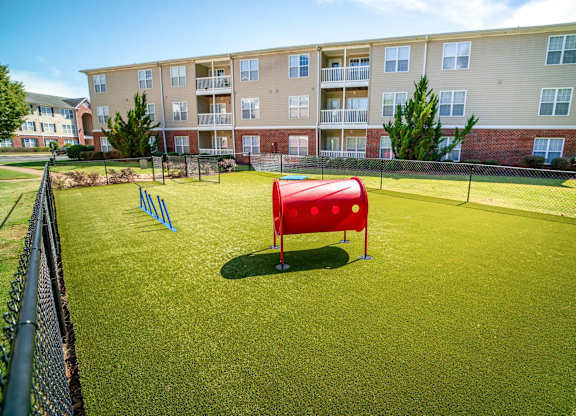 A red sand bucket sits on a green field in front of apartment buildings.