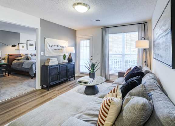 A living room with a grey sofa and a bedroom with a bed and a nightstand in the background.